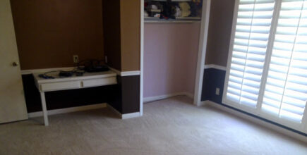 Spacious room with beige carpet, a white desk, and brown-painted walls with natural light from shuttered windows.