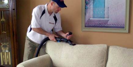A professional cleaner using a vacuum to clean upholstery on a beige sofa in a living room.