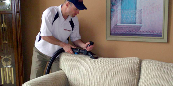 A professional cleaner using a vacuum to clean upholstery on a beige sofa in a living room.